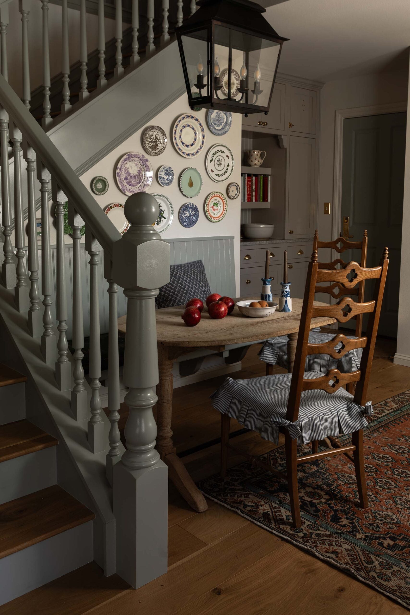 Oho Interiors-Carriage House-Wood Dining Table with Antique Chairs Candelabra Pendant Light Plate Collection on Wall of Spindle Staircase